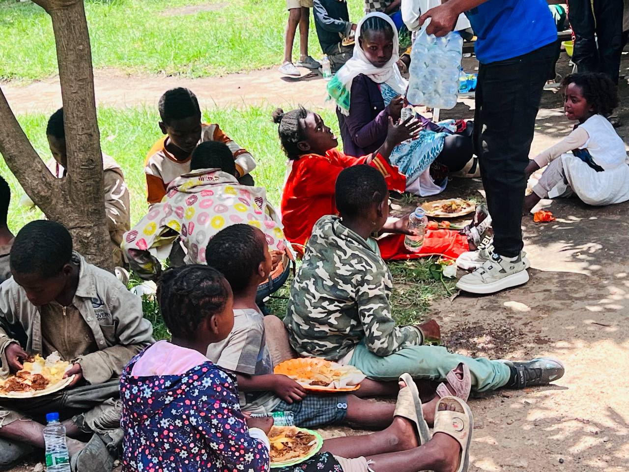 Children sitting under a tree eating a meal while a volunteer hands out water